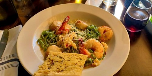 A plate of shrimp pasta with green noodles, topped with cheese and served with a slice of bread and a glass of red wine.