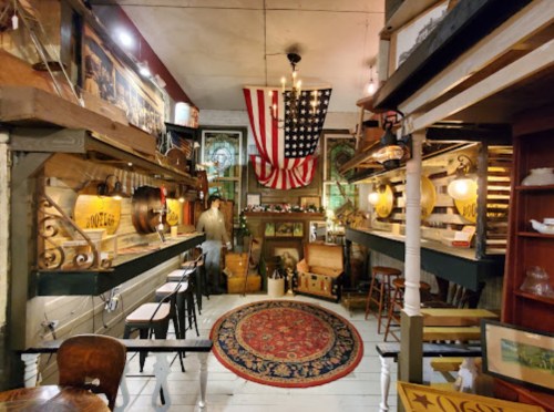 A vintage shop interior featuring wooden displays, an American flag, and various antiques on shelves.