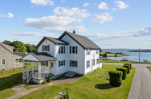 A white two-story house with a porch, overlooking a scenic waterfront and green landscape under a blue sky.