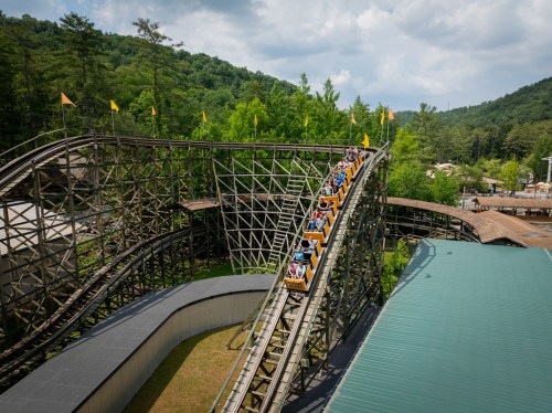 A wooden roller coaster with riders going down a steep curve, surrounded by green trees and a cloudy sky.