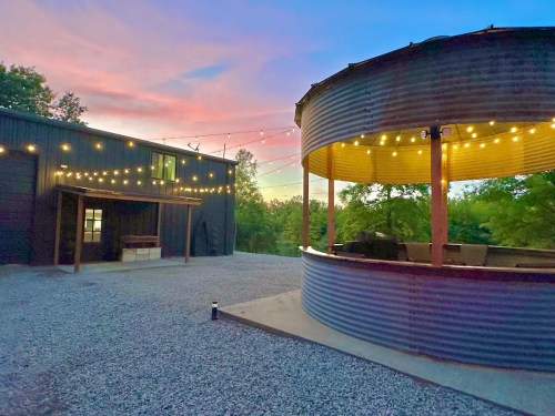 A rustic outdoor space with string lights, a circular bar, and a modern building against a colorful sunset sky.
