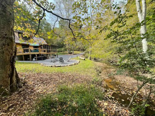 A serene cabin by a creek, surrounded by trees and autumn leaves, with a patio and seating area.