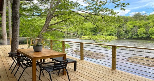 A wooden deck with a table and chairs overlooks a serene river surrounded by lush green trees.