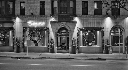 Black and white image of a restaurant facade decorated with wreaths and festive lights, set on a city street.