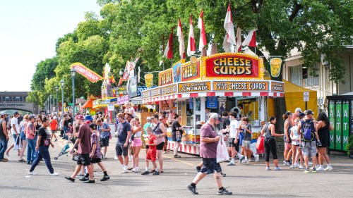A bustling fair scene with food stalls, including cheese curds, and crowds enjoying the festivities under sunny skies.
