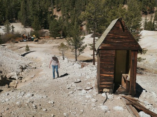 A person walks down a rocky path near an old wooden structure in a forested area. Trees and distant vehicles are visible.