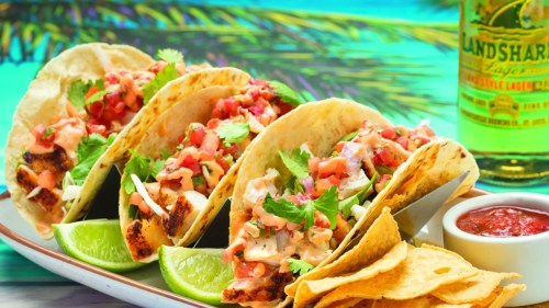 A plate of tacos topped with pico de gallo and cilantro, served with lime wedges, tortilla chips, and salsa.