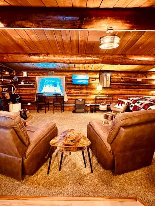 Cozy log cabin interior with two armchairs, a wooden table, and a view of a TV and window. Warm, inviting atmosphere.