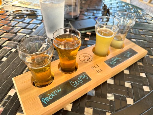 A wooden flight paddle with four small glasses of beer, labeled "Andrew," "Gypsy," "Harry," and "Me," on a table.
