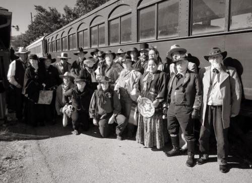 A large group of people dressed in Western attire poses in front of a vintage train, captured in black and white.