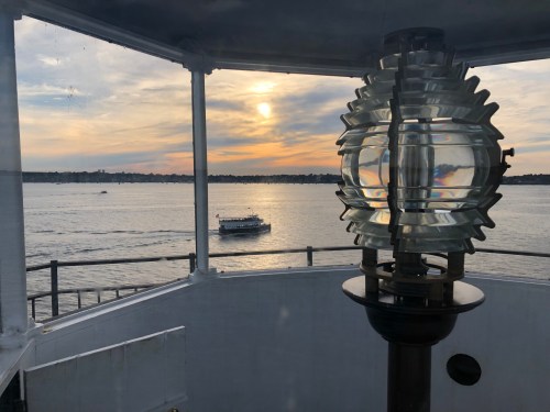View from a lighthouse with a glass lantern, overlooking a calm sea and a boat against a sunset sky.
