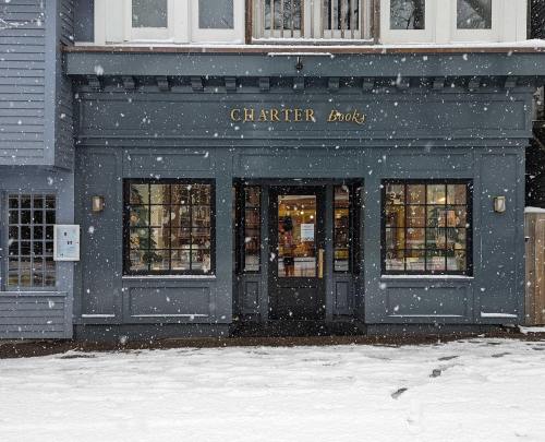 A cozy bookstore named "Charter Books" with large windows, surrounded by falling snow and a snowy ground.