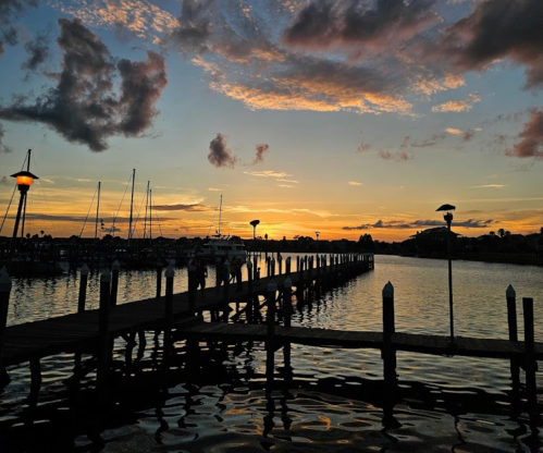 A serene sunset over a marina, with a wooden pier extending into calm waters and boats moored nearby.