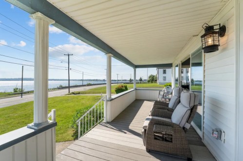 A cozy porch with two chairs overlooks a scenic view of a road and water under a clear blue sky.