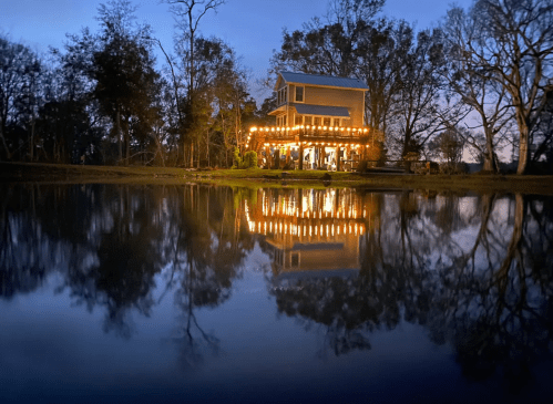 A charming house with string lights reflects in a calm pond at dusk, surrounded by trees.