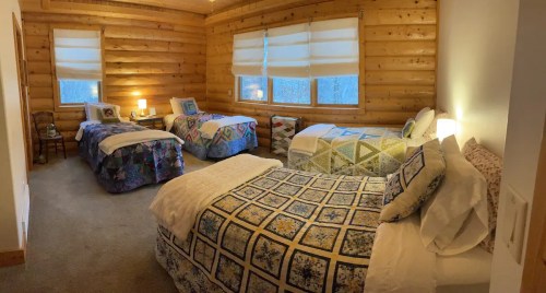 Cozy cabin bedroom with three beds, wooden walls, and large windows letting in natural light.