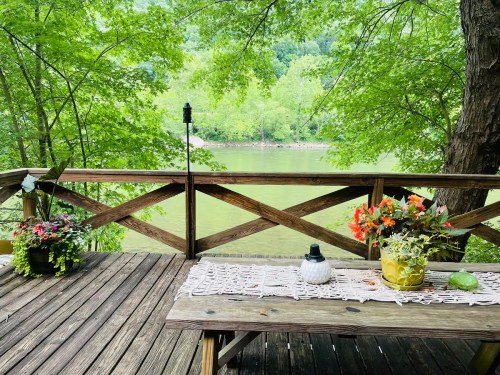 A wooden deck overlooking a serene river, adorned with flowers and a decorative table setting under lush green trees.
