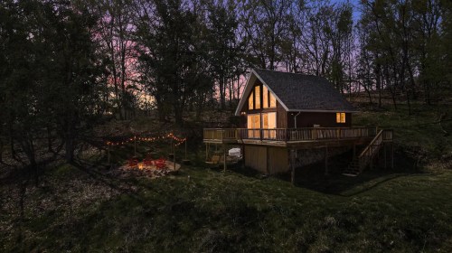 A cozy cabin with a deck, surrounded by trees, illuminated at dusk with a fire pit nearby.
