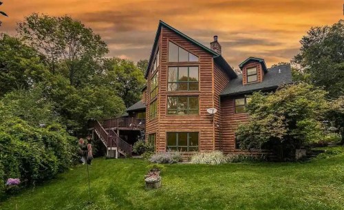 A large log cabin home surrounded by greenery, with a vibrant sunset sky in the background.
