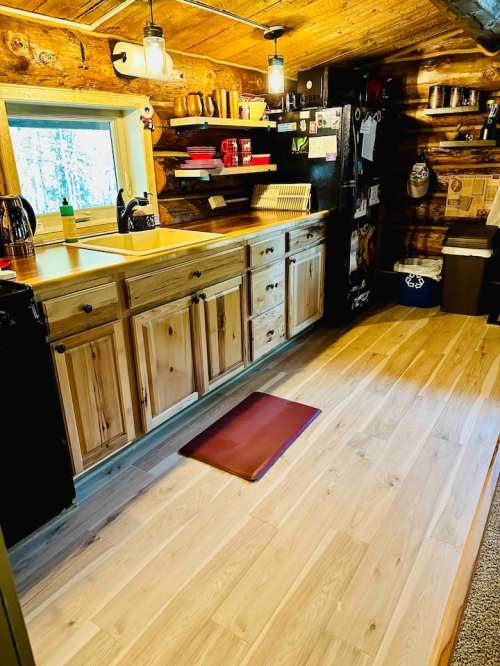 Cozy log cabin kitchen with wooden cabinets, a sink, and a mat on the floor, illuminated by warm overhead lights.