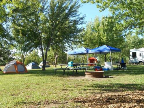 A campsite with orange tents, blue canopies, picnic tables, and a fire pit, surrounded by green trees and grass.