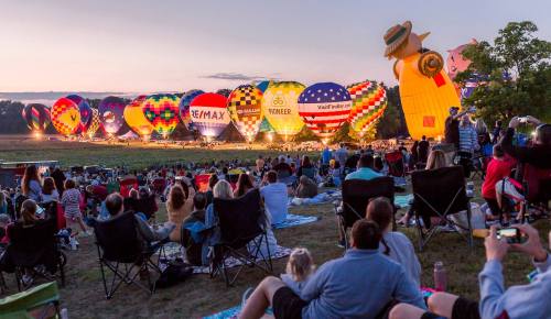 A crowd watches colorful hot air balloons illuminated at dusk during a festival, with blankets and chairs on the grass.