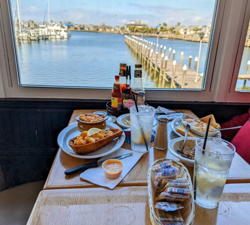A table with various seafood dishes and drinks, overlooking a scenic waterfront view with a dock.
