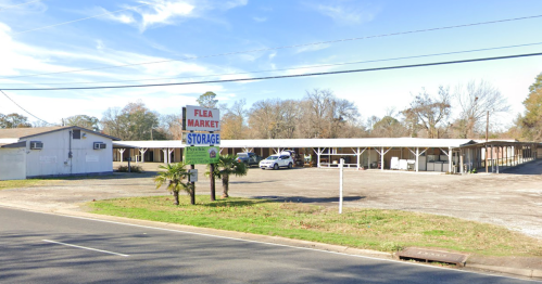 A flea market and storage facility with a sign, surrounded by trees and a gravel parking lot.
