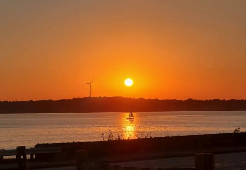 A serene sunset over a calm waterway, with a sailboat and a wind turbine silhouetted against the orange sky.
