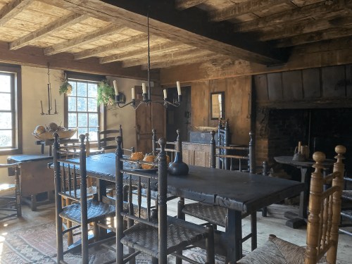 A rustic dining room with a large wooden table, chairs, a chandelier, and a fireplace, featuring warm, natural lighting.