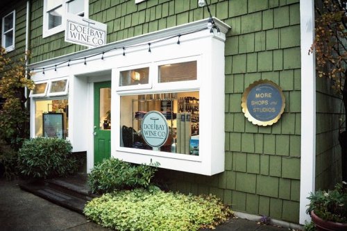 Exterior of Doe Bay Wine Co. featuring a green door, large windows, and a sign for more shops and studios nearby.