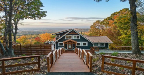 A modern house with a wooden walkway, surrounded by colorful autumn trees and a scenic view in the background.