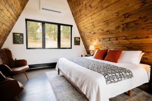 Cozy A-frame bedroom with wooden walls, a large window, a bed with decorative pillows, and a chair.