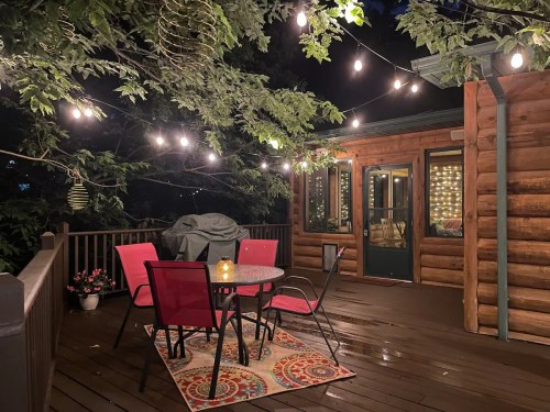 A cozy outdoor deck with a round table, red chairs, and string lights, surrounded by trees and a warm ambiance.