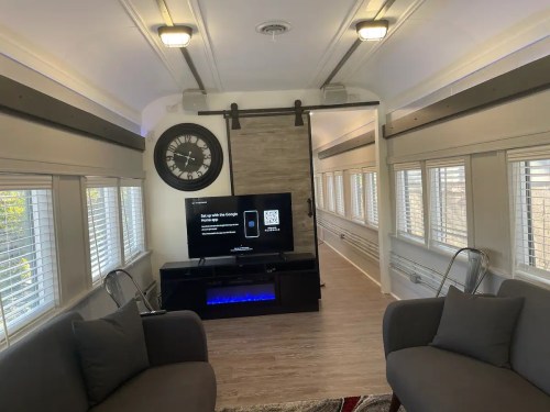Interior of a modern, well-lit room with a TV, seating area, and large windows, featuring a clock on the wall.