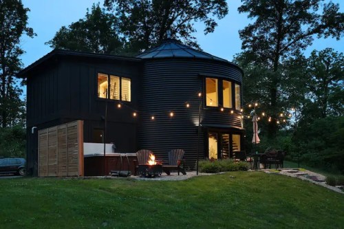 A modern round black house at dusk, with string lights, a fire pit, and a hot tub in a serene outdoor setting.