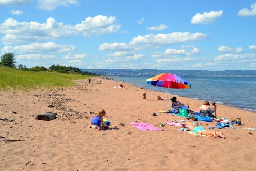 A sandy beach scene with families enjoying the sun under colorful umbrellas and playing near the water.