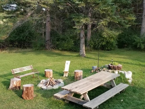 A serene outdoor space with a picnic table, benches, and a fire pit surrounded by trees and greenery.