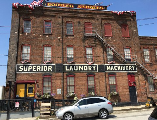 A brick building with "Bootleg Antiques" and "Superior Laundry Machinery" signs, featuring a car parked in front.