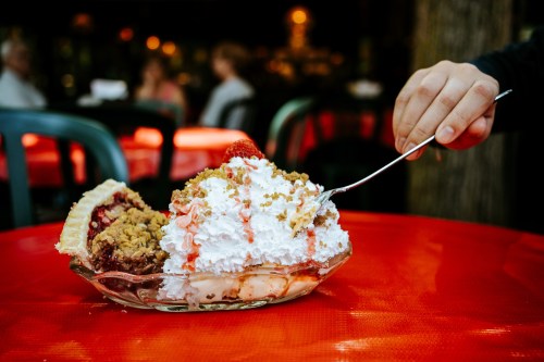 A hand holds a spoon over a large dessert with ice cream, whipped cream, pie, and strawberry sauce on a red table.