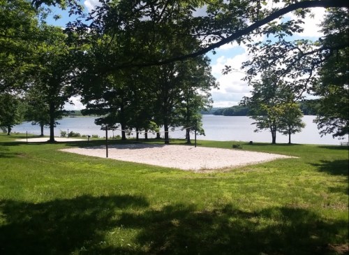 A sandy volleyball court by a lake, surrounded by trees and a clear blue sky.