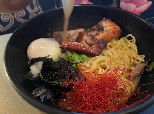 A bowl of ramen with noodles, sliced meat, a soft-boiled egg, seaweed, green onions, and red threads on top.