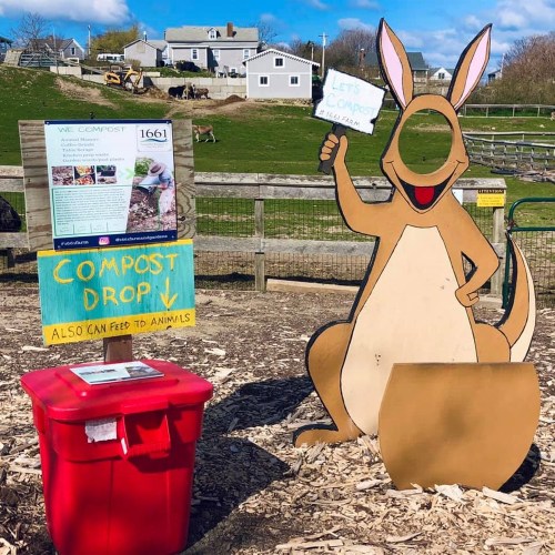 A cartoon kangaroo holds a sign promoting composting next to a red compost bin and an informational board.