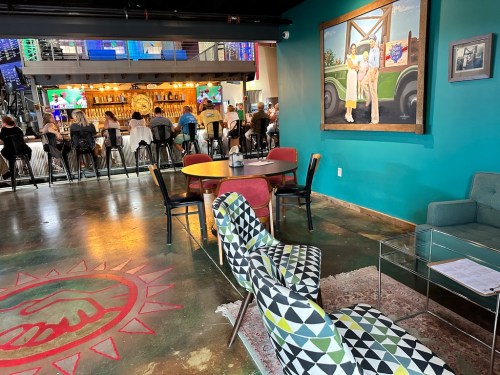 A vibrant interior featuring colorful seating, a bar area, and a large painting of children by a green tractor.