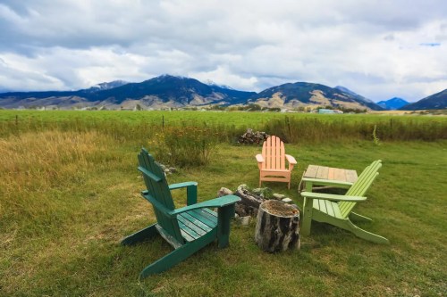 A serene outdoor scene with colorful chairs around a fire pit, set against a backdrop of mountains and grassy fields.