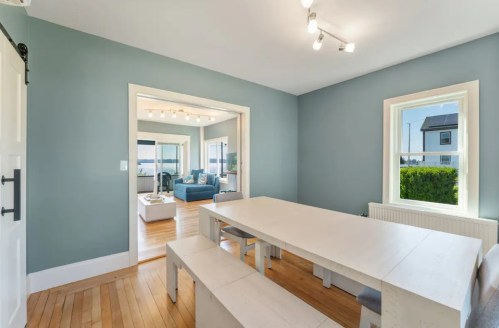 Bright dining area with a white table and benches, leading to a cozy living room with large windows and blue decor.