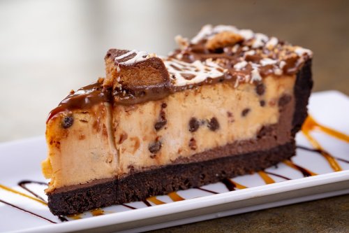 A slice of chocolate chip cookie dough cheesecake on a white plate, drizzled with caramel and chocolate sauce.