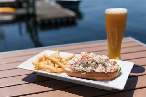 A lobster roll topped with seafood salad, served with fries and a glass of beer, set against a waterfront backdrop.