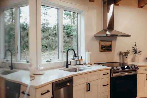 Modern kitchen with white cabinets, stainless steel appliances, and large windows overlooking greenery.