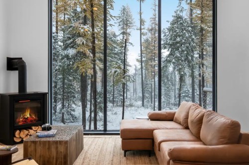 Cozy living room with a brown sofa, fireplace, and large windows showcasing a snowy forest landscape.